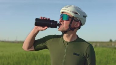 Bisiklet kasklı ve gözlüklü neşeli genç adam gün batımını izlerken spor şişesinden içiyor. Gevşek yol sürücüsü egzersiz yaptıktan sonra hızlı iyileşme için sıvı dengesini sağlıyor.