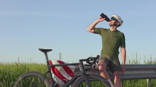 身穿运动服的运动员坐在路障上 靠着田园背景喝水 在夏天的艰苦训练之后 骑自行车的人非常享受旅行瓶子里的饮料 — 图库视频影像
