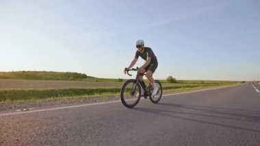 Açık yol bisikleti süren sporcunun yaz günbatımında kaldırımlı otoyolda güçlü bir şekilde pedal çevirmesi. Dinamik yoğunlukta bisiklet eğitimi almış deneyimli sporcu..