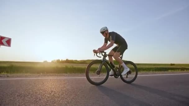 ロードバイクの完全装備のレーサーは フィールドバック上のロードウェイに沿って高速で運動ライドを行います 日没の忙しいトレーニングスケジュールに保つタイトフィットな服のスポーティな男性サイクリスト — ストック動画
