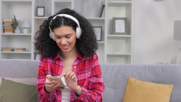 ハッピーな表情の女性がスマートフォンでメッセージを入力しながら 室内でワイヤレスヘッドフォンを介して音楽を楽しんでいます 現代技術を使用してアフリカ系アメリカ人女性の時間を過ごす — ストック動画