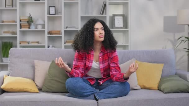 若いアフリカ系アメリカ人女性は 快適なソファーに座っている間 自宅でヨガを練習しています リラックスした女性の目を閉じて 屋内で瞑想します バランスとマインドフルネスの概念 — ストック動画