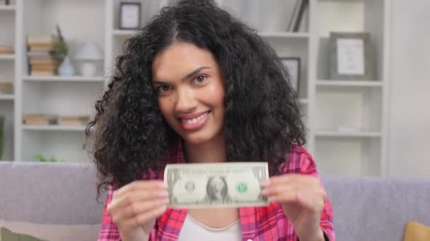 一个令人愉快的非洲裔美国女人的画像 她展示着一美元钞票 直视着镜头 快乐的女士手握现金 坐在舒适柔软的沙发上 坐在私人住宅里 — 图库视频影像