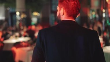 Güzel görünümlü, siyah şık takım elbiseli bir beyefendinin şık restoranlarda konuklarla birlikte yuvarlak masaların arasında güvenle yürüdüğü arka plan görüntüsü. Yeni evliler için erkek düğün sunucusu gösterisi.