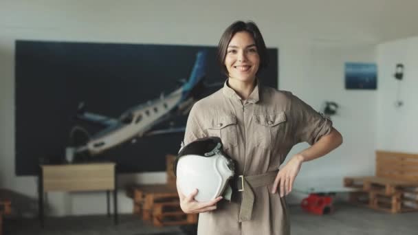 スタジオに立ち カメラで笑顔でヘルメットを持つ魅力的な白人女性パイロットの肖像画 飛行機で大きな画像の背景にポーズし 親指を上げる陽気な女性 — ストック動画