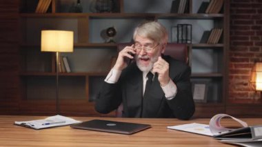 Gözlüklü, resmi takım elbiseli, yönetici koltuğunda otururken duygusal olarak akıllı telefondan konuşan yaşlı bir erkek. Ofisteki dijital teknolojileri kullanarak hissedarlara bağlanan deneyimli bir işletme sahibi.
