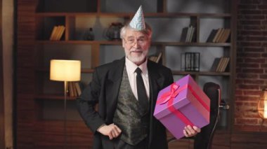 Doğum günü konisi şapkalı kıdemli işadamının portresi lüks bir ofiste dururken büyük bir hediyeyle poz veriyor. Gözlüklü genel müdür yeni şirket projesinin başarıyla başlamasını kutluyor.