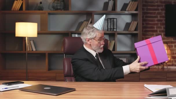 オフィスのエグゼクティブデスクに置いている間 贈り物を観察する誕生日の円錐形帽子の興奮した老人の側面の景色 職場で創業した企業を記念する正式な服装の陽気な社員 — ストック動画