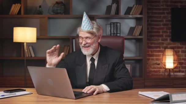 オフィスのラップトップでビデオ通話アプリを介して祝福を受ける紙コーン帽子の幸せな老人のビジネスマネージャー 同僚からの挨拶や願いに感謝の気持ちを表現する笑顔のプロフェッショナル — ストック動画