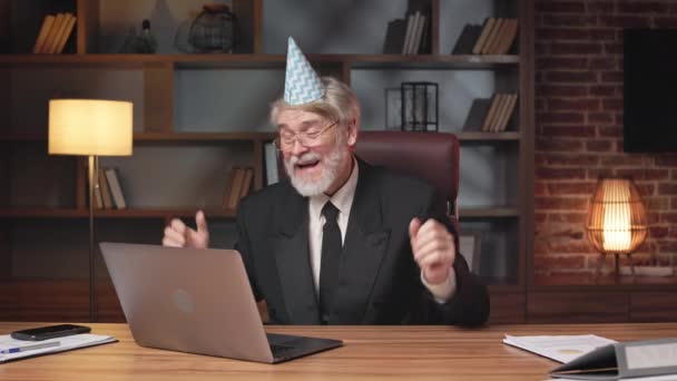 兴奋的商人大约60岁 穿着正式的服装 头戴生日帽 在工作场所的笔记本电脑上举行虚拟会议 快乐的胡子创业者在网上享受成功创业的赞美 — 图库视频影像