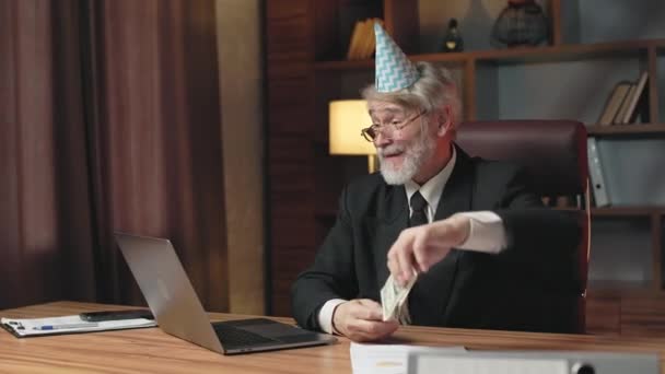 灰色头发的男性戴着眼镜 身穿西服 头戴生日帽 一边看着笔记本电脑屏幕 一边把纸币抛向空中 处理工作中的商业项目财务失败的年长雇员感到不安 — 图库视频影像