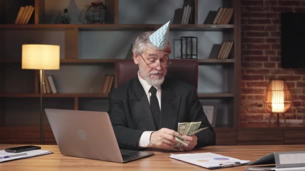 オフィスに木製の机に置く前に 紙の円錐形の帽子で不快な年上の男 職場でお金の問題に苦労しながらストレスを感じるビジネスマン — ストック動画