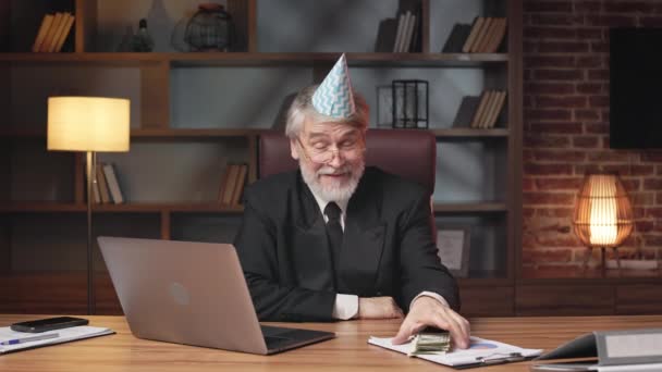 坐在笔记本电脑后面工作时 戴着派对帽的嘲讽的老年人把堆积如山的钞票放在写字台上 白发苍苍的白种人 穿着正式服装 在办公室里不给钱 — 图库视频影像