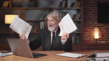 Resmi takım elbiseli mutlu yaşlı bir adam dizüstü bilgisayarlı web kamerasıyla online tartışmalar yaparken kağıt kayıtlarını havada tutuyor. Gözlüklü neşeli çalışan iş yerindeki proje onayı için heyecanlanıyor.