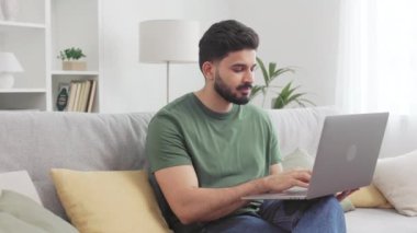 Çekici genç adam evdeki kablosuz bilgisayarda çalışırken evdeki interneti kullanarak rahat koltukta oturuyor. Neşeli gündelik giysiler içinde Hindu erkek serbest yazar. Mesafeli işten zevk alıyor..