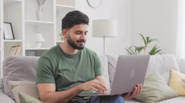 Gri rahat koltukta otururken portatif dizüstü bilgisayarda çalışırken yüzünde hafif bir gülümseme olan yakışıklı Hintli adam. Serbest çalışma ve aygıtlar konsepti.