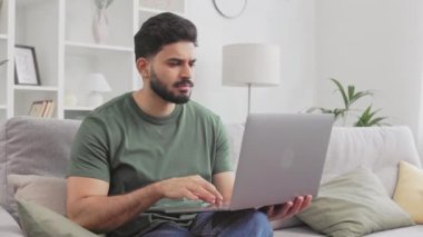 Rahat giysiler içinde, çenesini kaşıyan ve kanepede otururken dizüstü bilgisayarda düşünceli bir şekilde görünen yakışıklı Hintli bir adam. Erkek serbest çalışan evde soru çözme fikirleri arıyor..