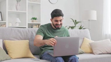 Güzel haberleri el hareketleriyle kutlayan sıradan giyinmiş yakışıklı Hintli adam kanepede oturup dizüstü bilgisayar ekranına bakıyor. İnsan kavramı, teknoloji ve başarı.