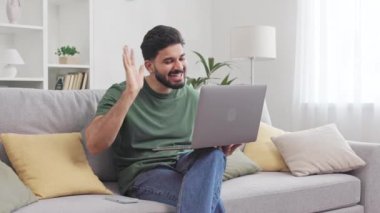 Modern bilgisayardaki video konferansında karşılama için el sallayan gülümseyen sakallı adam. Çekici Hintli erkek gündelik kıyafetlerle kanepede oturuyor ve online iletişimin tadını çıkarıyor..