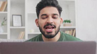 Evdeki video görüşmesi için kablosuz bilgisayarı kullanan yeşil tişört giymiş yakışıklı bir adam. Genç Hintli adam uzaktan kumanda sırasında kanepede oturuyor ve online toplantı yapıyor..