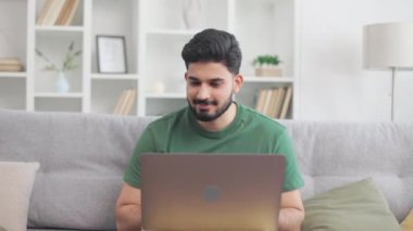 Koyu saçlı, sakallı, rahat koltukta oturan ve taşınabilir dizüstü bilgisayarda yazan Hintli bir adam. Çekici genç adam evde otururken uzaktan işten zevk alıyor..
