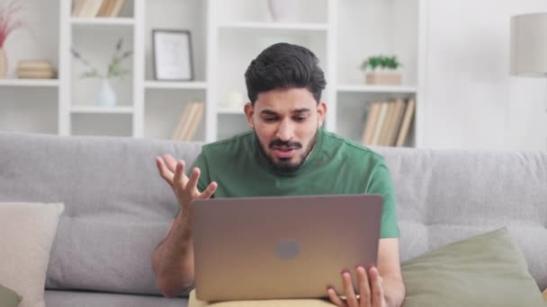 現代のラップトップでストレスを受けたビデオ会話中にヒンドゥーの男性が叫び ジェスチャーをしました 同僚とのオンラインコミュニケーションからの否定的な感情を持つソファに座っている男性フリーランサー — ストック動画