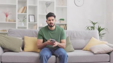 Evde mobil uygulama sorunu yaşayan sakallı Hintli bir adam kendini rahatsız hissediyor. Sıradan giyimli Hindu erkek gri koltukta oturuyor ve sinirleniyor..