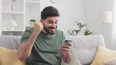 Modern akıllı telefonu elinde tutan tatmin olmuş Hintli adam internetten gelen harika haberlere hayran kalıyor. Duygusal sakallı erkek rahat bir evde kanepede otururken yumruğunu sıkarak zaferi kutluyor..