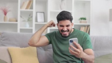 Gri kanepede rahat bir oturma odasında otururken, elinde cep telefonu tutan ve kazanan gibi el kol hareketi yapan, gündelik giysiler giymiş duygusal Hintli bir adam. Samimi tepki ve servet kavramı.