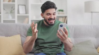 Modern cep telefonundan harika haberler alırken yumruğunu zevkle sıkan mutlu yakışıklı adam. Sakallı Hindu erkek evdeki kanepede dinleniyor ve gerçek mutluluğu kontrol edemiyor..