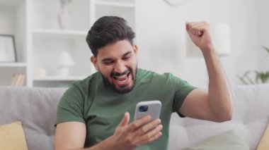 Yakışıklı sakallı adam modern akıllı telefondan harika haberler okuyarak duygusal olarak seviniyor. Hindu genç adam oturma odasında rahat bir kanepede otururken el hareketleriyle kutlama yapıyor..