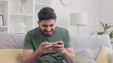 Kanepeye oturup video oyunları oynamak için modern akıllı telefonu kullanan sıradan giyinen heyecanlı sakallı adam. Hintli erkek oyuncu rahat koltukta dinleniyor ve kaybettiği halde sonuçtan memnun değil..