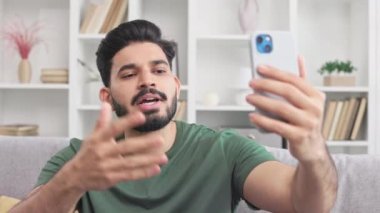 Modern cep telefonuyla video konferansı sırasında parmaklarıyla konuşan ciddi Hindu erkek. Oturma odasındaki rahat koltukta oturan ve internetten iletişim kuran genç adam..