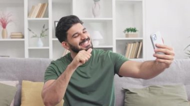 Gülümseyen yakışıklı adam başparmak yapıyor, tamam mı? Ve modern akıllı telefondan selfie çekerken barış işaretleri. Milenyum çocuğu gri koltukta oturuyor ve sosyal medya için yeni içerikler hazırlıyor..