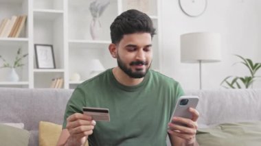 Mutlu Hintli adam rahat koltukta oturuyor ve online alışveriş için kredi kartıyla modern akıllı telefonu kullanıyor. Çekici milenyum adamı internet siparişleri için uygun ödemenin keyfini çıkarıyor..