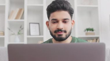 Sıradan giyinen sakallı genç adam evdeki uzaktan kumanda için kablosuz laptop kullanıyor. Yakışıklı Hindu adam gri koltukta oturuyor ve pozitif yüz ifadesiyle ekrana bakıyor..