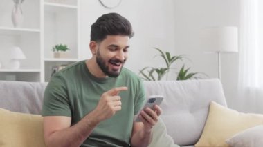 Yakışıklı Hintli adam kanepede oturuyor, ağzını açıyor ve şaşırmış bir yüz ifadesiyle akıllı telefon ekranına bakarken büyük gözler yapıyor. Beklenmedik haber ve tepki konsepti.