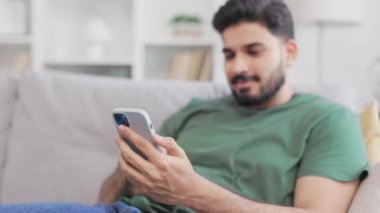 Gri koltukta rahat rahat oturan ve modern akıllı telefonu elinde tutan rahat ve çekici bir adam. Pozitif Hintli adam evde boş vakitlerini internette geçiriyor..