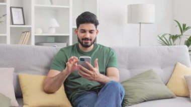 Gri kanepede otururken gündelik giyinmiş Hintli bir adam modern akıllı telefondan internete göz atıyor. Millennial 'lı genç adam elinde aletlerle rahat bir dairede dinleniyor..