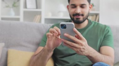Sakallı genç adam kişisel akıllı telefonuyla sosyal ağlara kayıyor ve evde dinlenirken gülümsüyor. Sıradan giyinmiş Hintli bir erkek kanepede oturuyor ve olumlu bakıyor..
