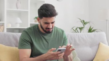 Hintli adam, gri koltukta tek başına otururken çalışmayan akıllı telefondan rahatsız oluyor. Koyu saçlı, yeşil tişörtlü genç adam evde modern aletlerle sorun yaşıyor..