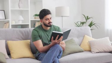 Kişisel dairede rahat bir kanepede dinlenirken mutlu sonla biten kitap okuyan pozitif Hintli adam. Yeşil tişört giyen yakışıklı erkek gelişimi için boş zaman harcıyor..