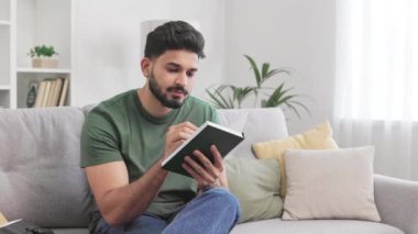 Pozitif Hindu erkek kişisel not defterine planlar yazarken dairedeki rahat koltukta iç mimari tasarım ile oturuyor. Günlük kıyafetler giymiş esmer adam not almak için kalem kullanıyor..
