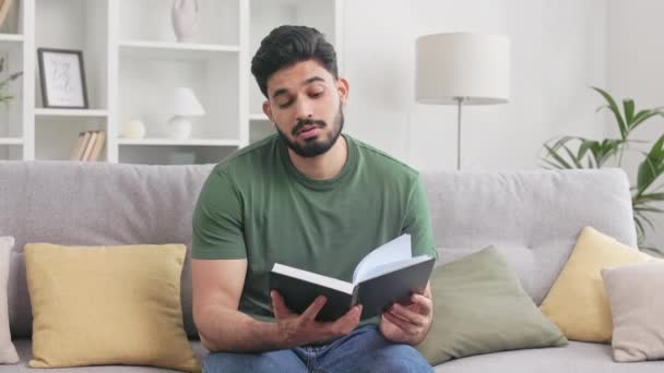 スマートフォン依存症の陽気なヒンドゥーの男性は文学を読むことに集中することはできません 満足した男は 本を置き 現代の携帯電話でウェブサイトをスクロールし始める — ストック動画