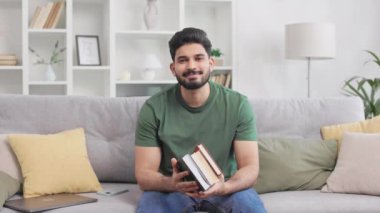 Rahat koltukta oturup kitap tutan, günlük giysiler giymiş pozitif Hintli öğrenci portresi. Koyu sakallı, ev ödevi yapan ve evde kalıp ders vermeye hazırlanan bir erkek..