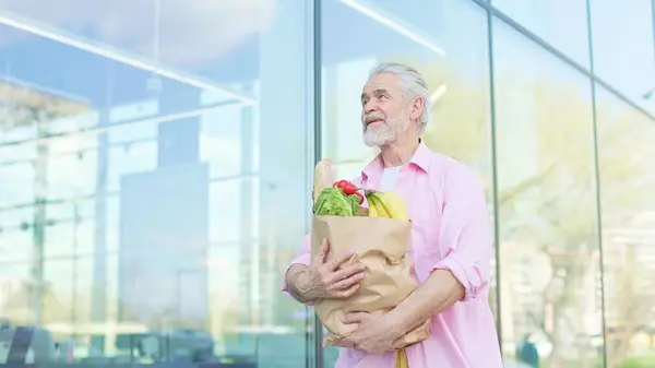 Beyaz, olgun bir erkek, panoramik camsı pencerelerle binanın yanında yürürken sağlıklı ürünlere sarılıyor. Sağlıklı yaşam tarzları ve insanlar kavramı.