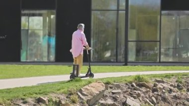 Günümüz şehir parkında elektrikli scooterla gezen gündelik giysiler giymiş yaşlı bir adam. Kentsel manzarada gezinmek için çevre dostu taşımayı seçerek çevreyi korumaya teşvik eden erkek..