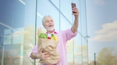 Olgun bir erkek cep telefonuyla selfie çekiyor. Arkaplanda da sağlıklı ürünler var. Yetişkin beyaz erkek blogcu sağlıklı yaşam tarzı hakkında vaaz veriyor ve kameraya yemek gösteriyor..