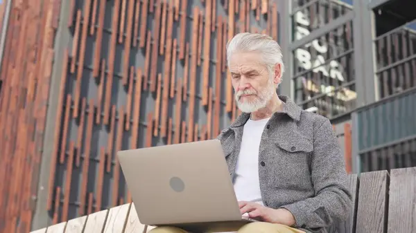 Yetişkin erkek, arka plandaki yerleşik binaların üzerindeki ahşap bankta dinlenirken kablosuz bilgisayarla çalışıyor. Gülümseyen beyaz adam dışarıda dinlenirken modern aletlere yazı yazıyor..
