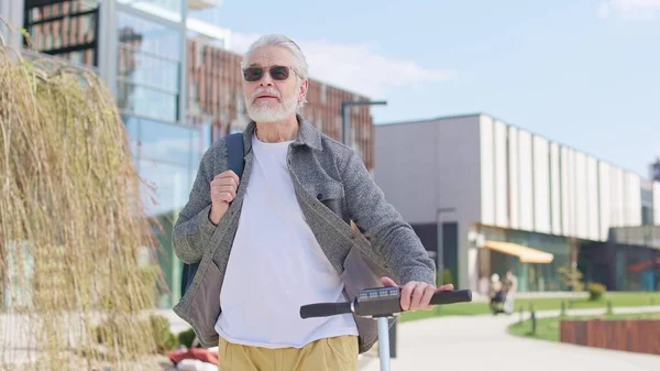Güneşli bir günde patikayı takip ederken sırt çantalı, elektrikli scooter kullanan çağdaş bir adam. Serin güneş gözlüklü, aktif bir yaşam tarzı olan ve şehirdeki eğilimlere ayak uyduran gri saçlı emekli bir erkek..